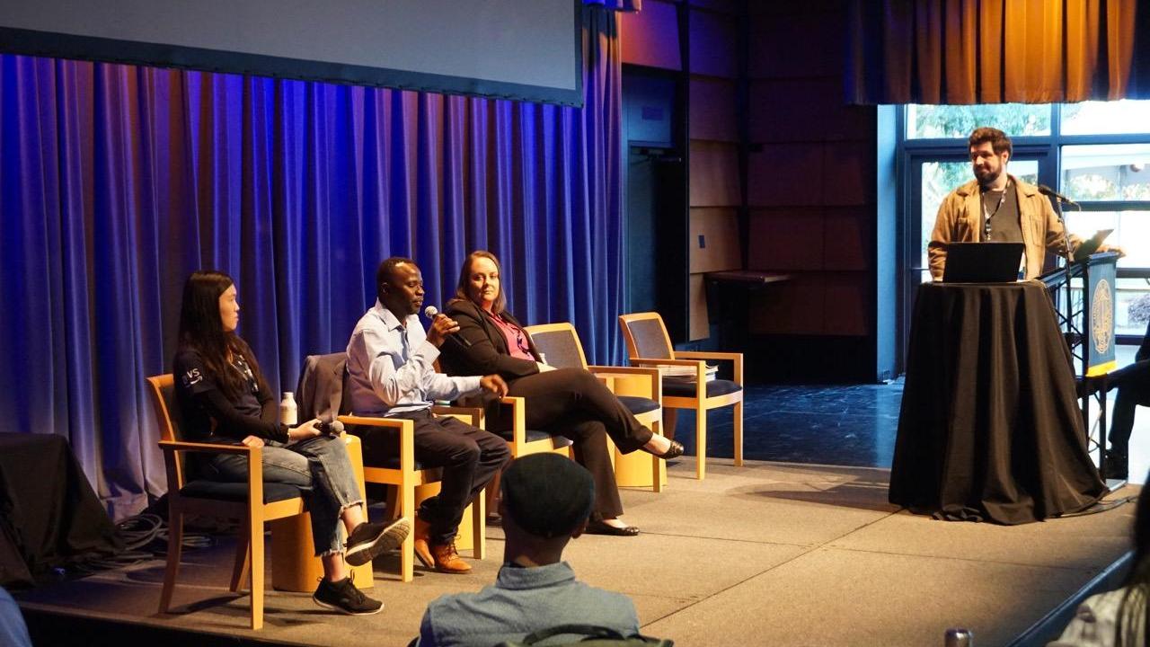 A panel at UC Tech 2024. Three people are sitting with mics, as one of them talks into a mic. The moderator looks at them from the side.