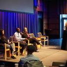 A panel at UC Tech 2024. Three people are sitting with mics, as one of them talks into a mic. The moderator looks at them from the side.